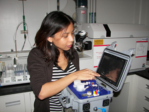 PNNL scientist Wassana Yantasee demonstrates the compact and field-portable biomonitoring device (foreground), which produces rapid, accurate results equivalent to state-of-the-art mass spectrometry systems (back) from small fluid samples.