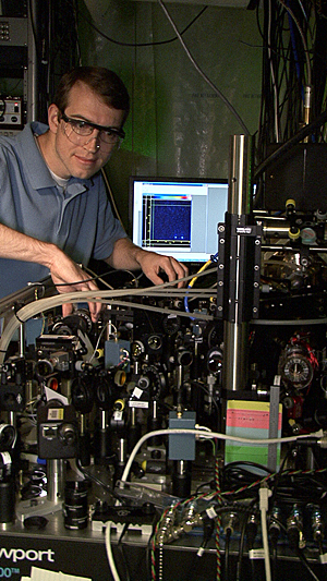 NIST postdoctoral researcher David Hanneke at the laser table used to demonstrate the first universal programmable processor for a potential quantum computer. A pair of beryllium ions (charged atoms) that hold information in the processor are trapped inside the cylinder at the lower right. A colorized image of the two ions is displayed on the monitor in the background.

Credit: J. Burrus/NIST
