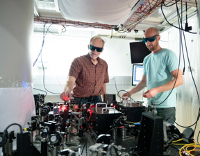 NIST physicists John Bollinger (left) and Matt Affolter adjust the laser and optics array used to trap and probe beryllium ions in the large magnetic chamber (white pillar at left). The ion crystal may help detect mysterious dark matter.

CREDIT
Jacobson/NIST