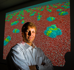 Sandia researcher Matt Lane stands before computer simulations of 2-nm. gold particles too small to measure experimentally. The particles aggregate to produce cigar-shaped objects that prefer to sit at the waters surface. Red represents oxygen, blue carbon, white hydrogen, yellow the sulfur coating. The gold particles are not modeled directly. (Photo by Randy Montoya)