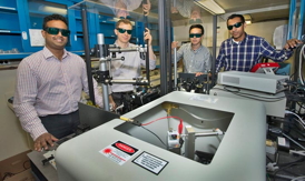Click on the image to download a high-resolution version. Nanditha Dissanayake, Matthew Eisaman, Yutong Pang, and Ahsan Ashraf display the setup used to track the flow of electrons through the photoactive layer of organic solar cells. The red and black wires in the box in the foreground (also shown in close-up) are connected to a solar cell that is in contact with a prism. The prism guides laser light through the cell in a range of specific directions to gain precise information about how electrons flow.